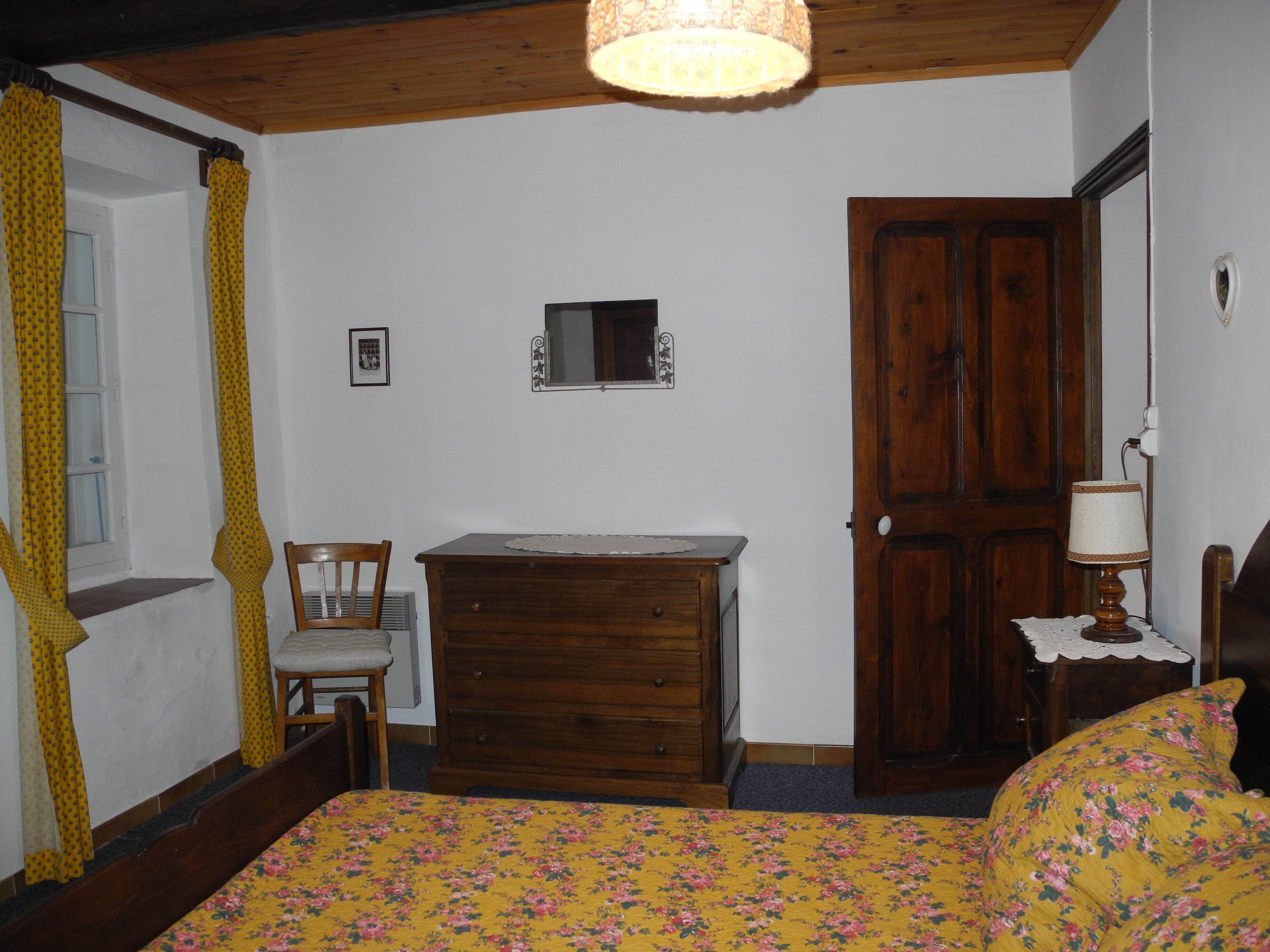 A bedroom with a wooden dresser, a chair, a bed with a floral bedspread, a wooden door, and a small bedside table with a lamp.