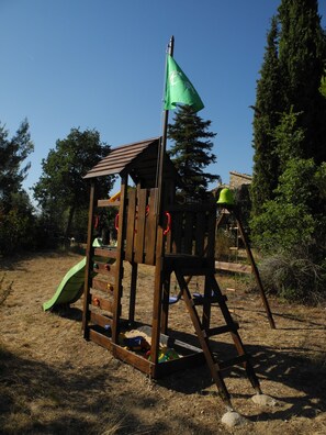 A wooden play structure with a slide and climbing area, surrounded by trees and dry grass.