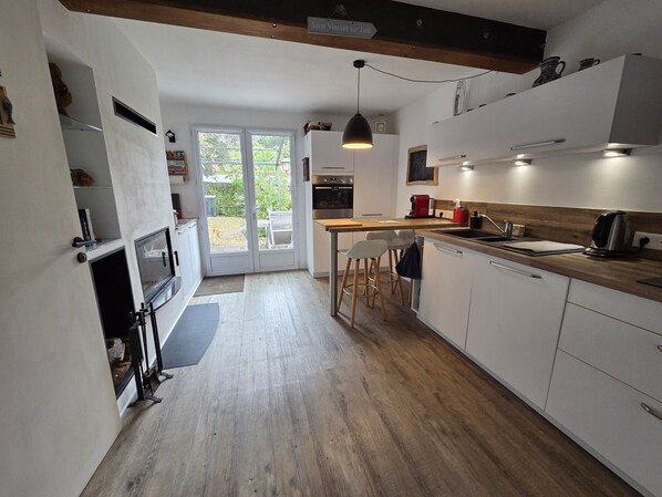 Une cuisine moderne avec un plancher en bois, des armoires blanches, un coin repas avec des tabourets et une vue sur l’extérieur grâce à des portes vitrées.