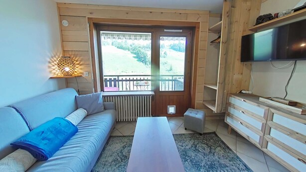 Ein gemütliches Wohnzimmer mit einer Couch, einem Couchtisch, einem Fernseher und Blick auf einen Garten durch ein großes Fenster.