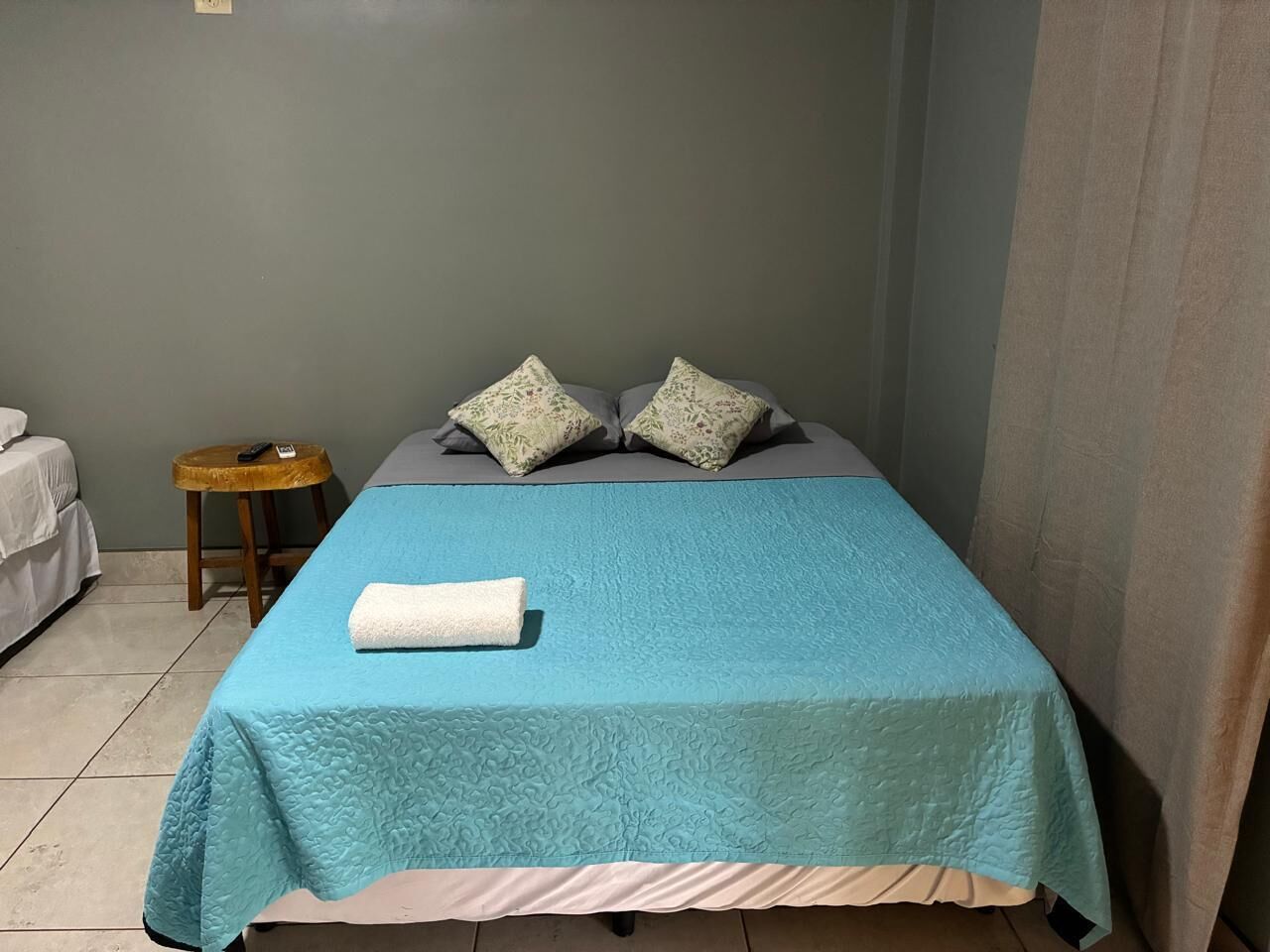 A single bed with a blue quilt and two pillows, a small wooden stool with a remote control, and a white towel on the bed.