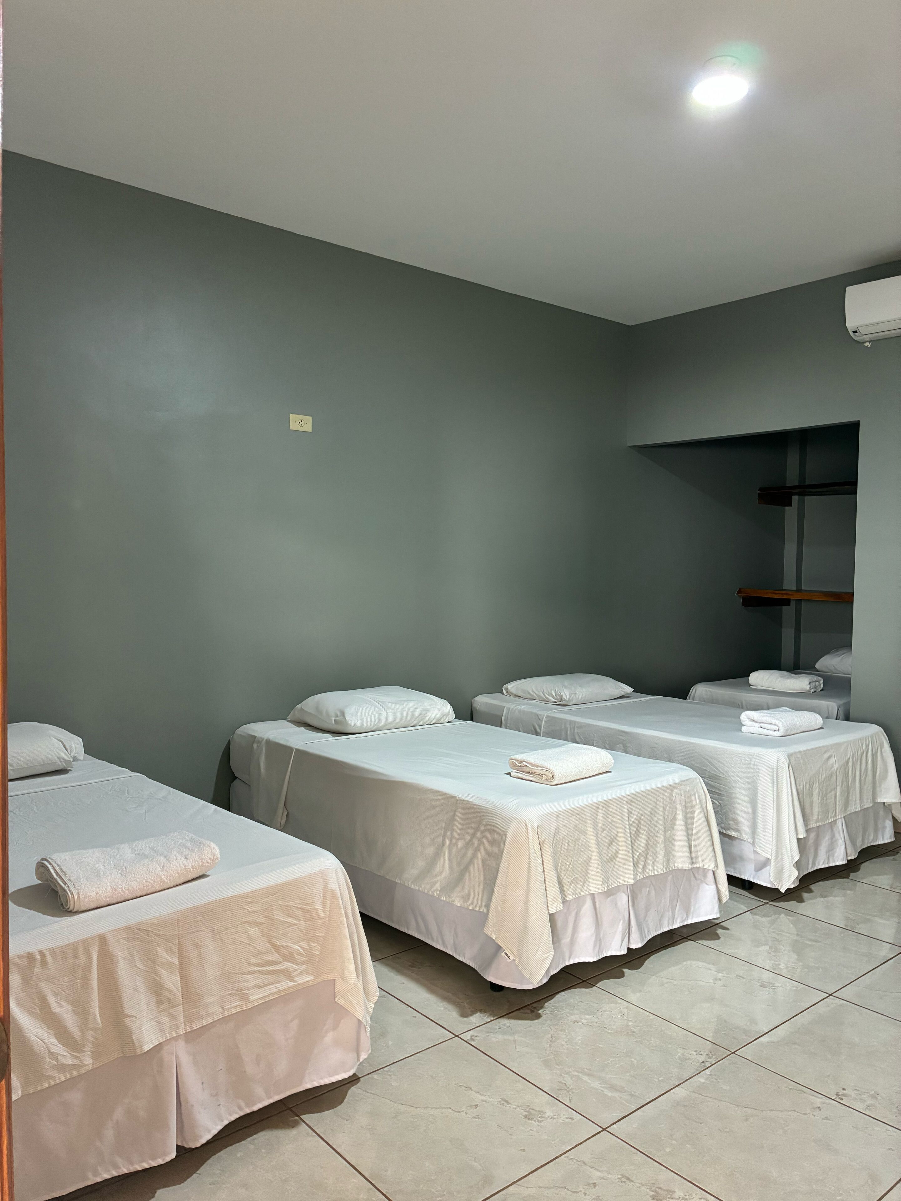 A room with four single beds, each with a white bedspread and a folded towel. The walls are painted a muted green, and there is a ceiling light.