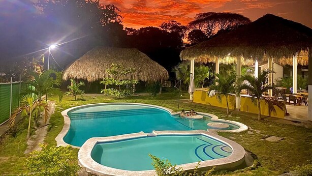 Une piscine, avec une structure à toit de chaume à proximité, entourée de verdure et surplombant un ciel nocturne parsemé de nuages.