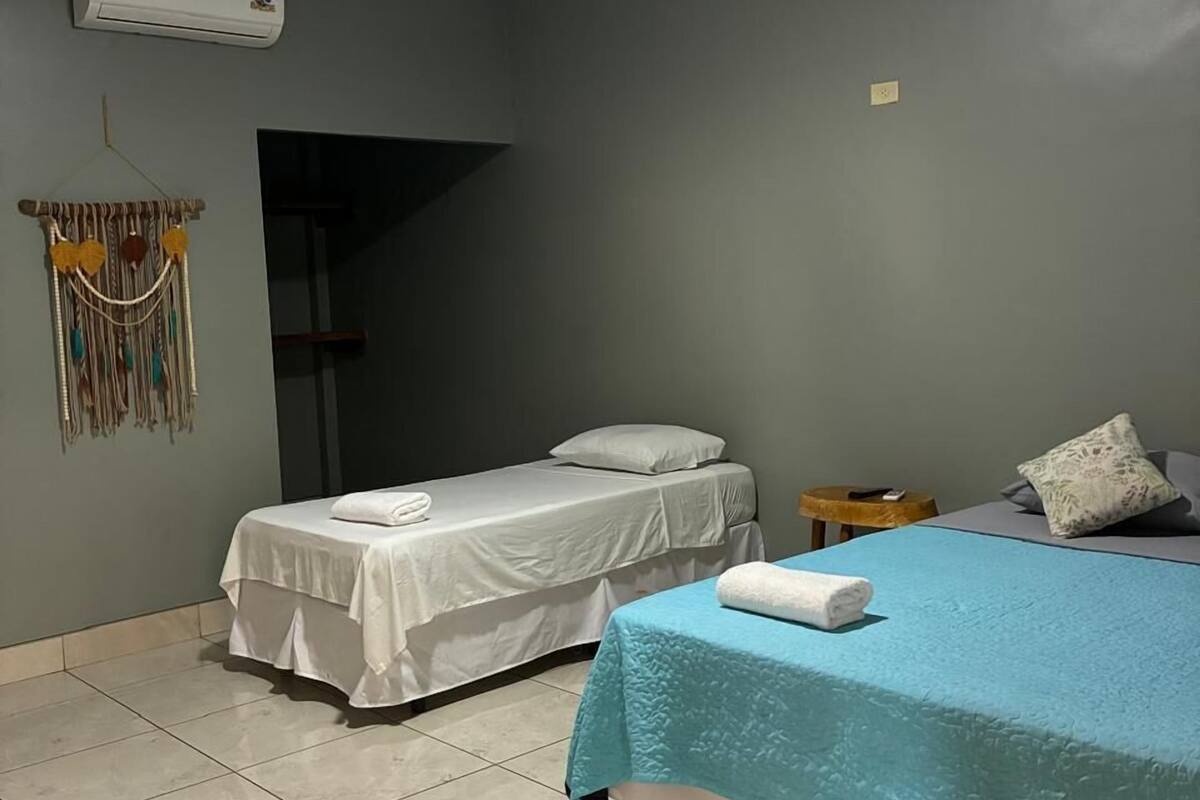 Ein Zimmer mit zwei Betten, eines mit blauer Bettdecke und das andere mit weißer Bettwäsche. An der Wand hängt eine Klimaanlage, ein kleiner runder Tisch und ein dekoratives Wandbild.