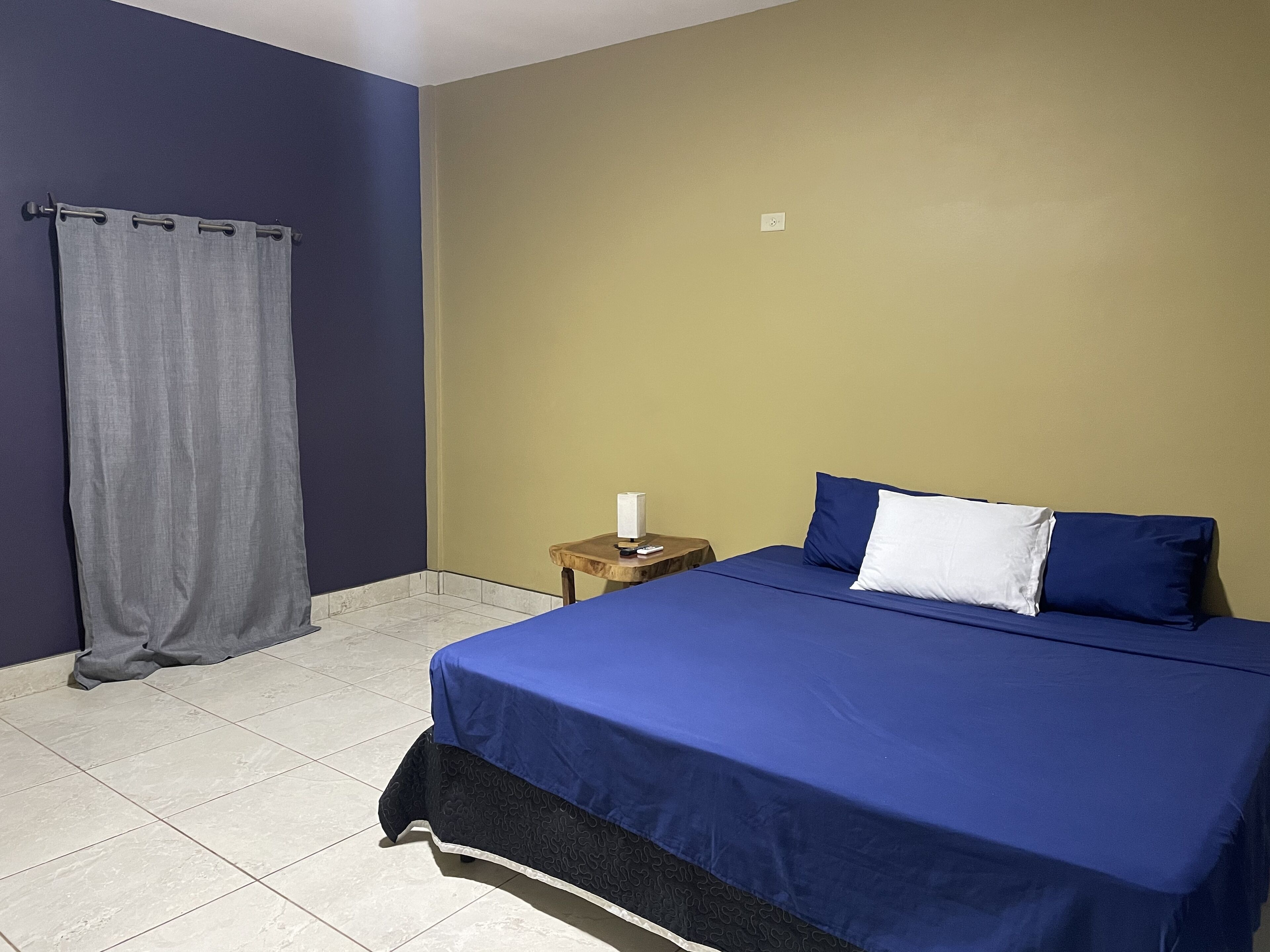 Un dormitorio con cama, mesita de noche, cortina y un interruptor de pared.