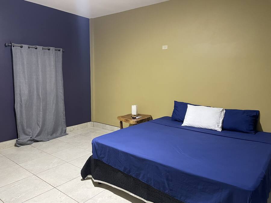 Un dormitorio con cama, mesita de noche, cortina y un interruptor de pared.