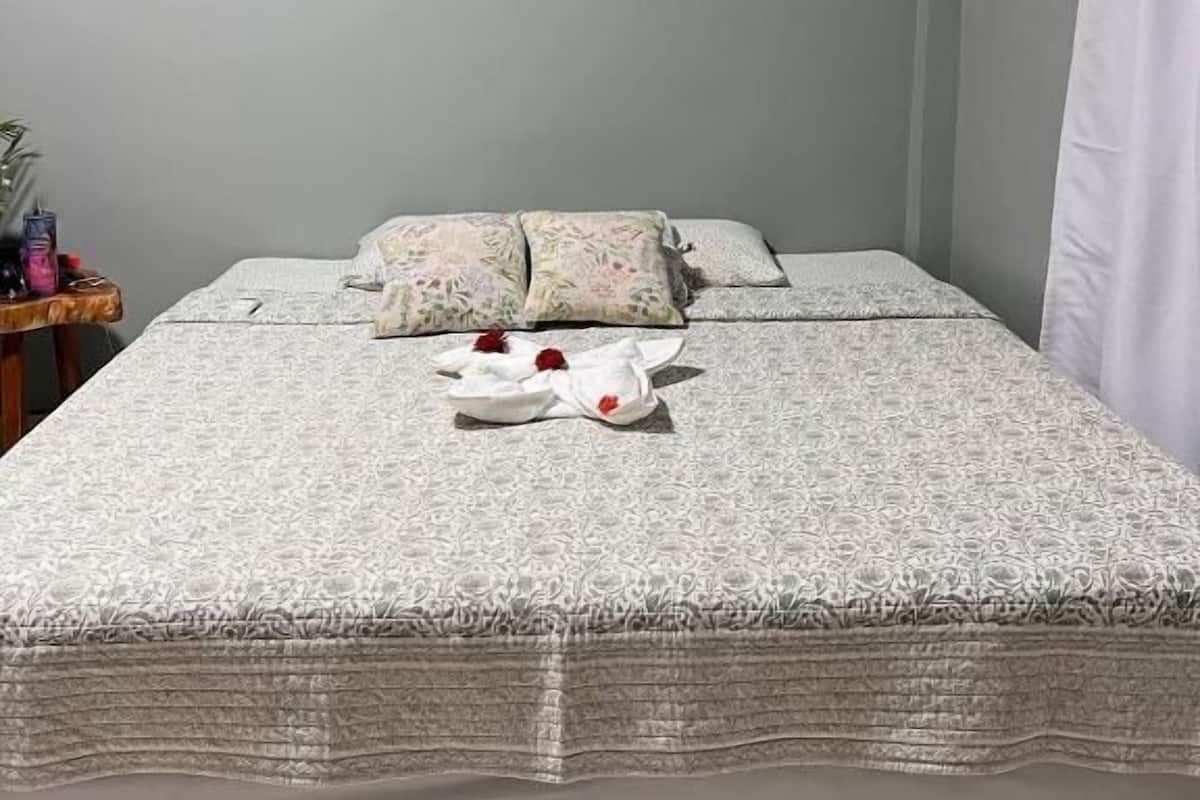 Ein ordentlich bezogenes Bett mit einer gemusterten Tagesdecke, ein Kissen mit floralem Dekor und ein Paar Slipper mit roten Blumen.