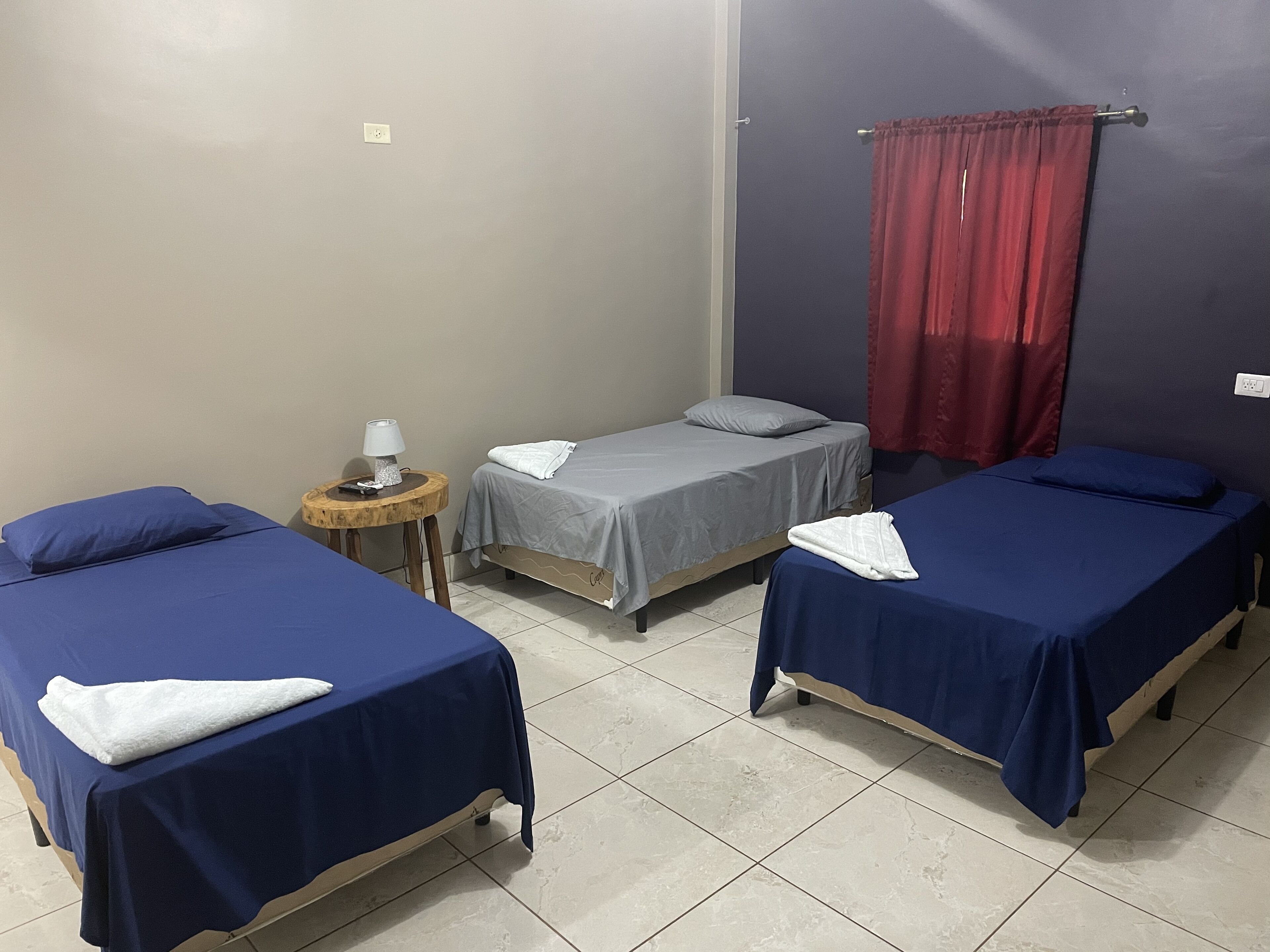 A room with three single beds, a small round table with a lamp, and a red curtain.