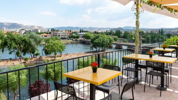 An outdoor terrace with tables and chairs, overlooking a river and a bridge, with buildings and trees in the background. - Lemon Cave Suites And Coffee House (Avanos)