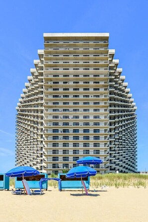 Un immeuble de grande hauteur doté de nombreux balcons, une plage de sable au premier plan et des parasols bleus.