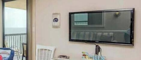 A room with a television mounted on the wall, a small white cabinet with books, a blue chair, and a window with a view.