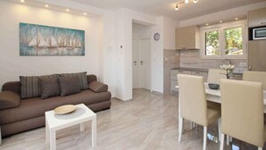 A modern living room with a sofa, a coffee table, and a dining area with chairs and a table. There is a painting on the wall and a clock on the door. - Holiday apartment, modernly furnished, only 300 m from the sea (Baška)