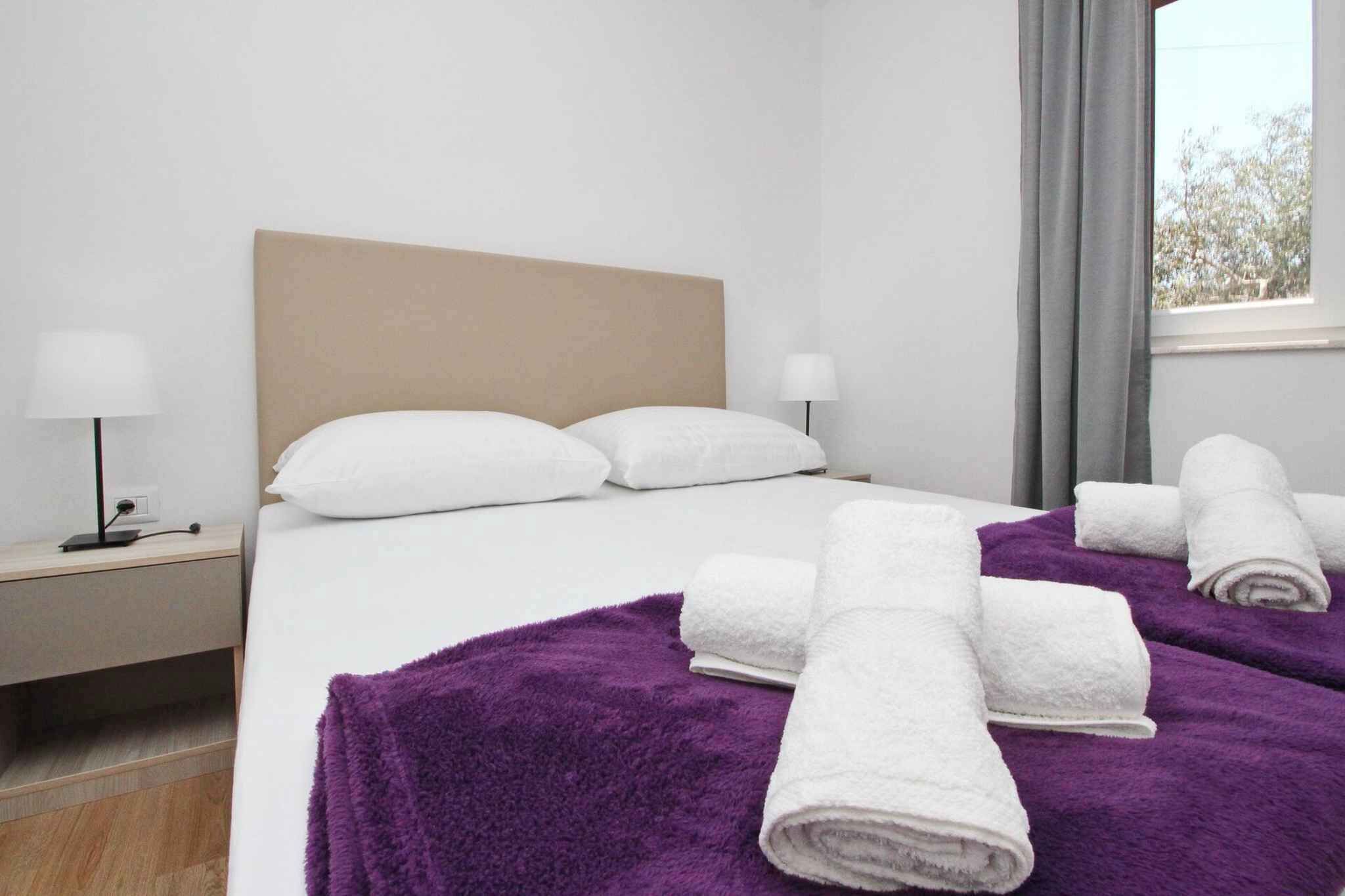 Una camera d'albergo con un letto, due comodini, una finestra con tende e una coperta viola sul letto.