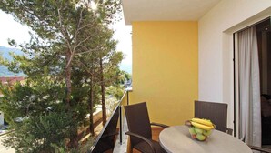 Un balcone con sedie in vimini e un tavolo, una ciotola di frutta e vista su alberi e palazzi.