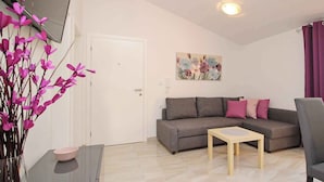 A living room with a grey sofa, a small wooden table, a vase with pink flowers, and a painting on the wall.