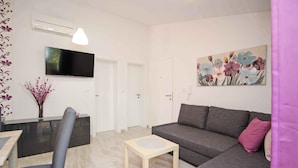 A modern living room with a grey sofa, a small wooden table, a TV mounted on the wall, and a floral painting.