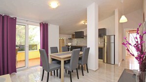 A modern dining area with a wooden table and chairs, a kitchen in the background, and a balcony with purple curtains.