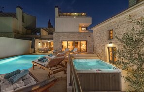 Et poolområde med liggestole og jacuzzi, omgivet af stengårde og en moderne bygning med balkon.