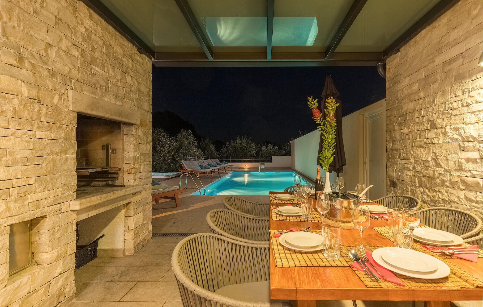An outdoor dining area with a pool, stone walls, wicker chairs, and a table set for a meal.