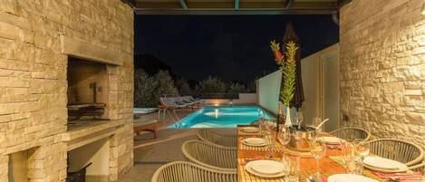An outdoor dining area with a pool, stone walls, wicker chairs, and a table set for a meal.