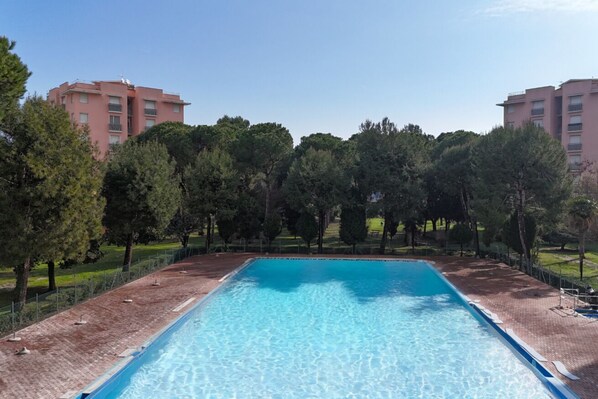 A rectangular swimming pool with clear blue water, surrounded by a brick deck and trees, with buildings in the background.