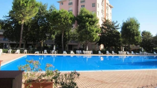 Una piscina con sillones de descanso, un edificio con balcones y árboles al fondo.