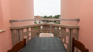 Un balcón con mesa y sillas, vistas a edificios y árboles, y una pared de color rosa.