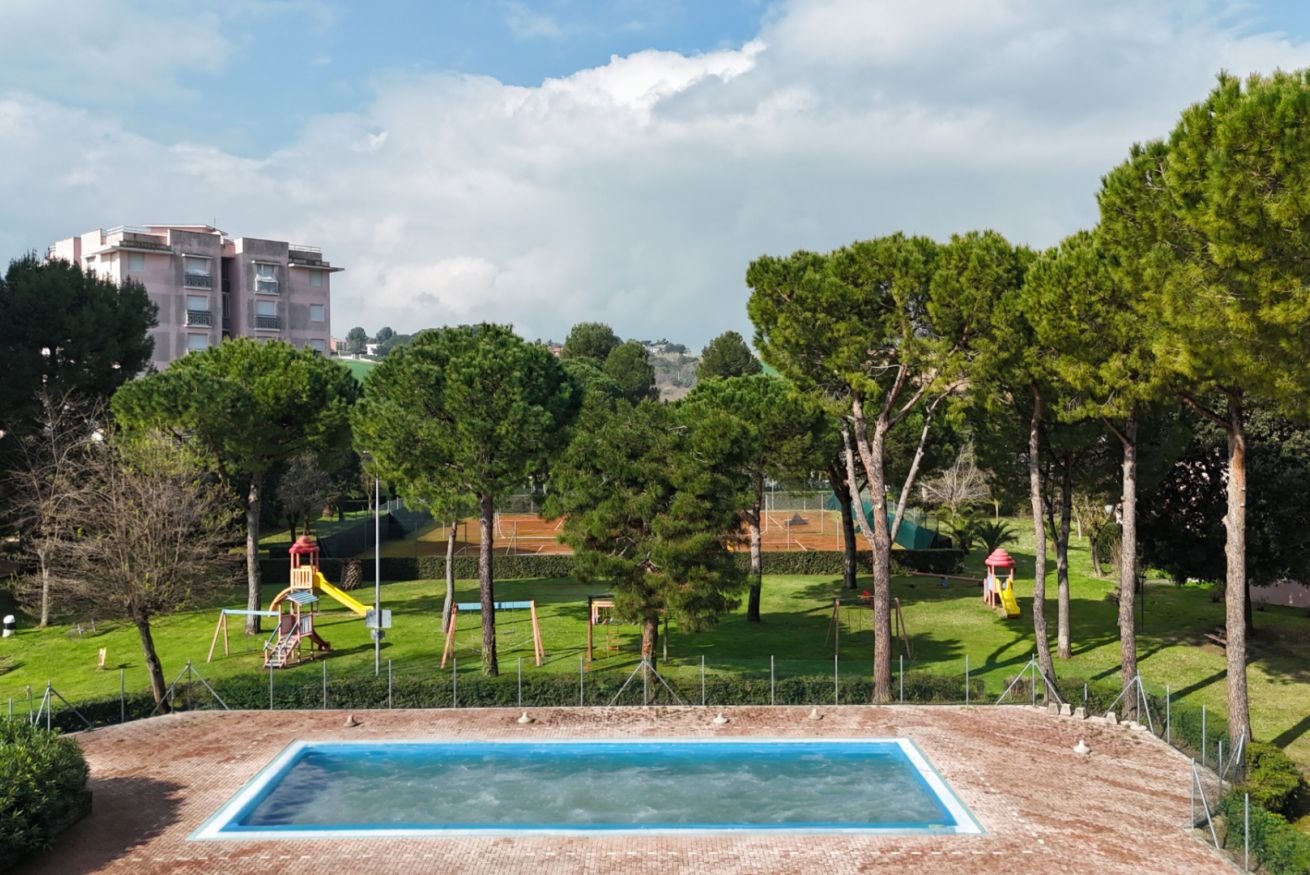 Ein Schwimmbecken, umgeben von einem eingezäunten Bereich mit Spielgeräten und Bäumen.
