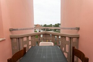 Un balcón con mesa y sillas, vistas a edificios y árboles, y una pared de color rosa.