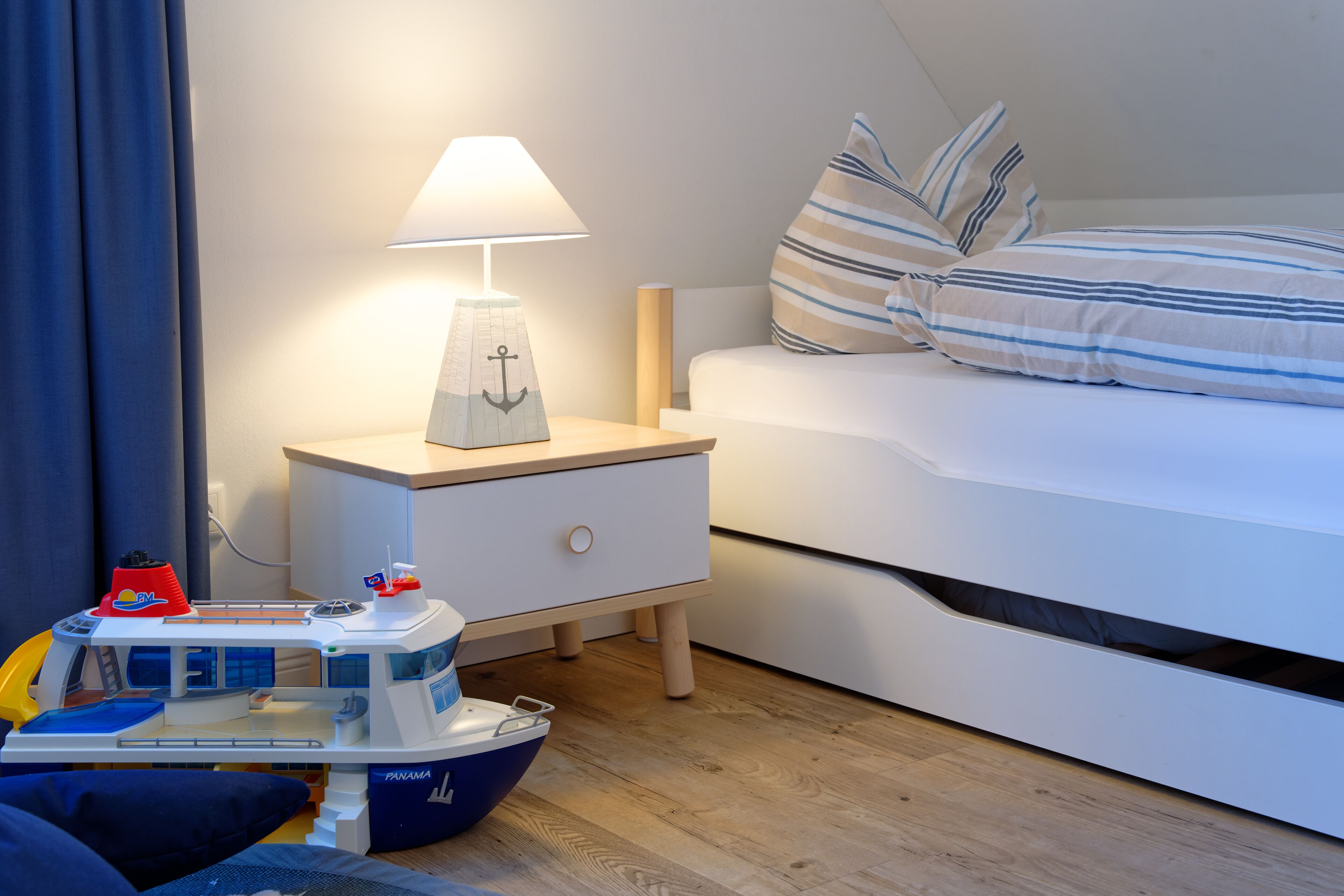 A hotel room with a bed, a nightstand with a lamp, a small desk, and a toy ship.