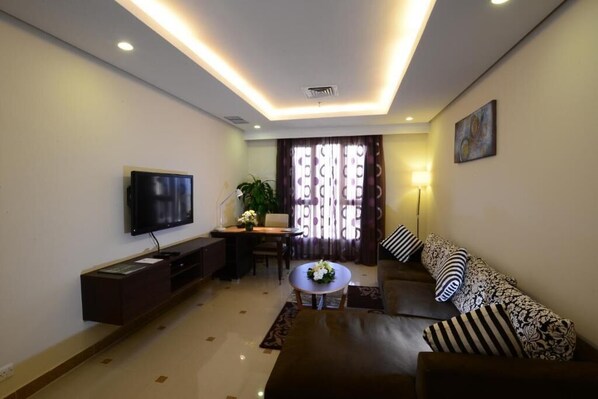 A living room with a flat-screen TV, a sofa with patterned cushions, a coffee table, and a dining area with a table and chairs.