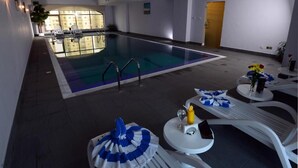 An indoor swimming pool with lounge chairs and tables, a wooden door, and a view of a building in the background. - The Venue Resident (Mahboula)