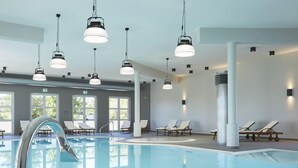 Sisätiloissa sijaitseva uima-allas, jossa on kirkasta vettä, ja sen ympärillä on rentoutumiseen sopivia aurinkotuoleja sekä modernit riippuvalaisimet.