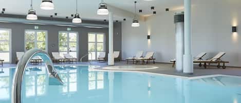 An indoor swimming pool with clear water, surrounded by lounge chairs and modern pendant lighting.