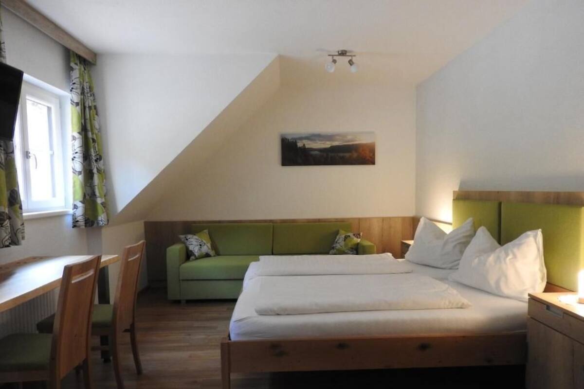 Une chambre avec un lit, un bureau et une chaise. Il y a une fenêtre avec des rideaux, un tableau au mur et un luminaire au plafond.