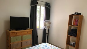 A bedroom with a bed, a TV on a wooden dresser, a standing fan, a window with curtains, and a shelf with toiletries. - Deluxe Apartment with Breakfast Included. (Villa Gral. Belgrano)