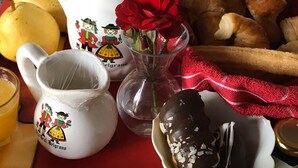 A breakfast setting with a teapot, mug, bread, a red rose, and a chocolate-covered pastry on a red tablecloth. - Deluxe Apartment with Breakfast Included. (Villa Gral. Belgrano)