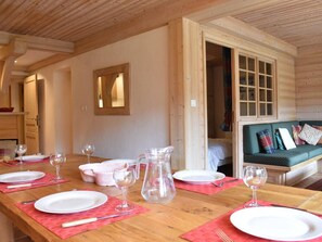 Une table de salle à manger en bois, mise pour quatre personnes, avec des assiettes, des verres et des couverts. On aperçoit, à l’arrière-plan, un banc en bois agrémenté de coussins.