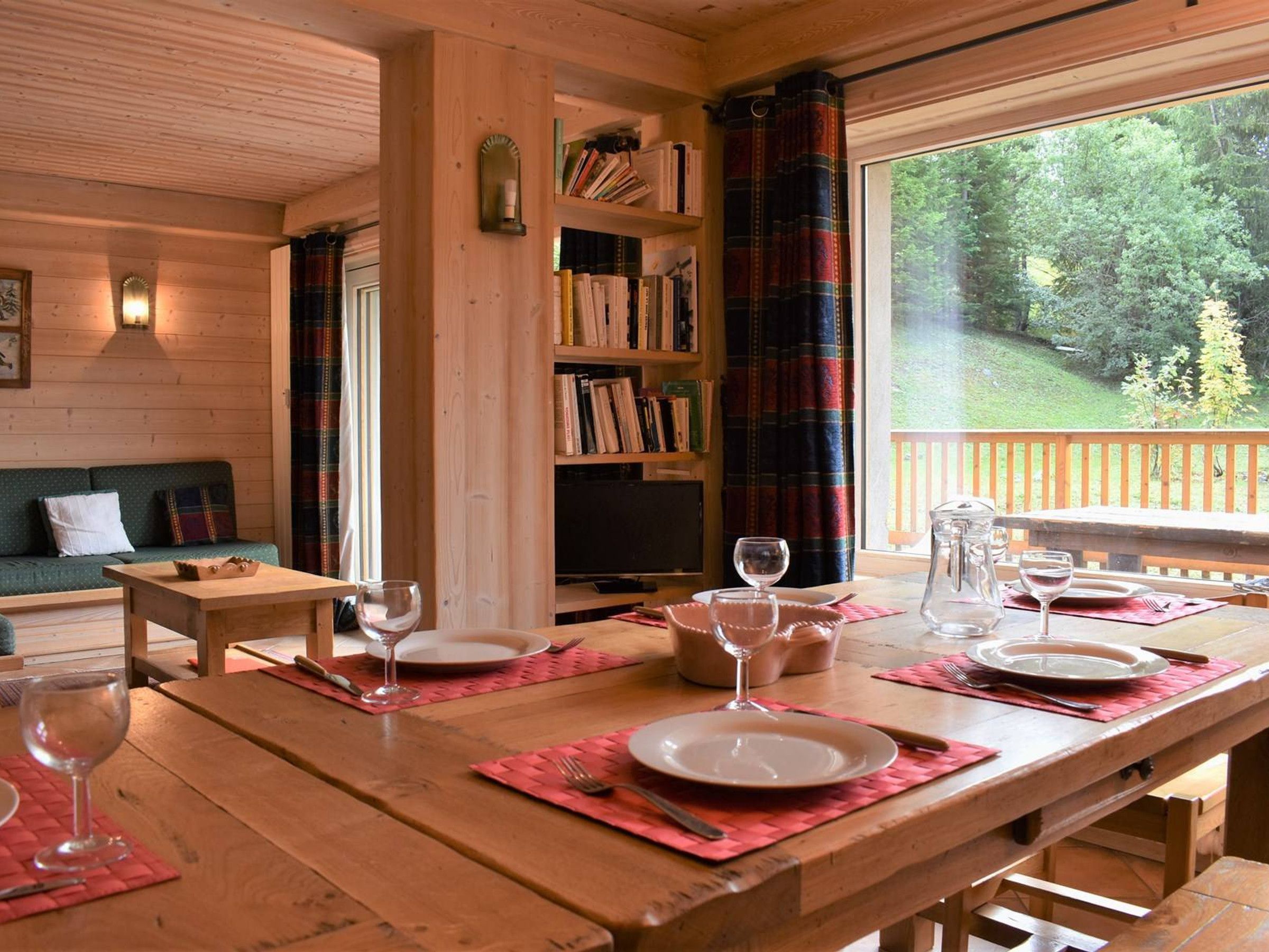 Una mesa de comedor de madera dispuesta con platos, vasos y cubiertos, una estantería con libros, un televisor y una vista de zona boscosa a través de una ventana.