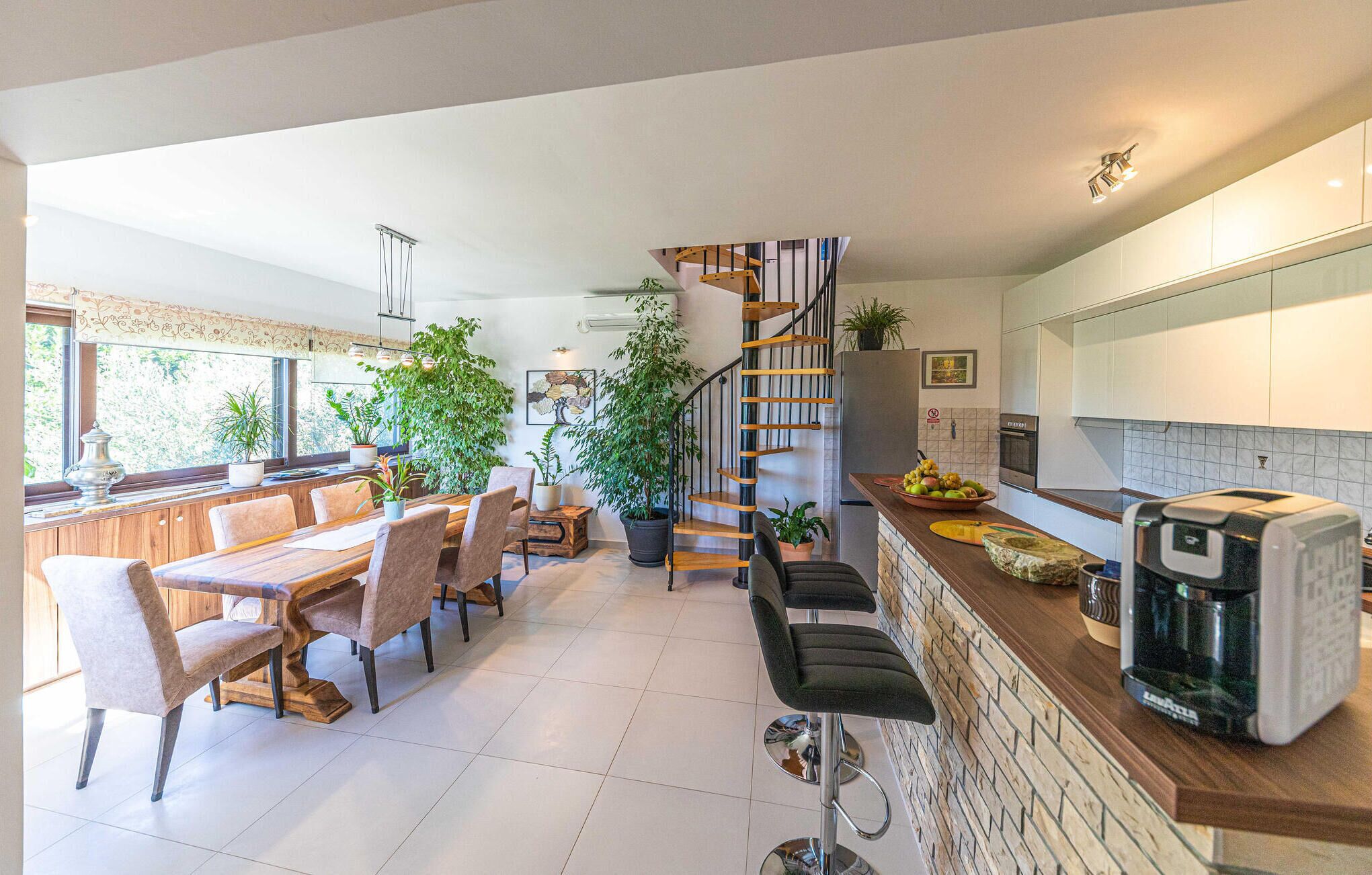 A modern kitchen with a wooden staircase, a dining area with a large table and chairs, and a kitchen counter with appliances and a coffee machine.