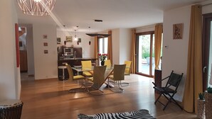A modern dining area with a glass table and yellow chairs, a kitchen with a microwave, and a view of the outdoors through large windows.