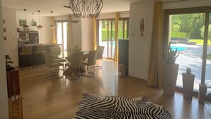 A modern living room with a glass dining table, comfortable chairs, a zebra-patterned rug, and a view of a swimming pool through large windows.
