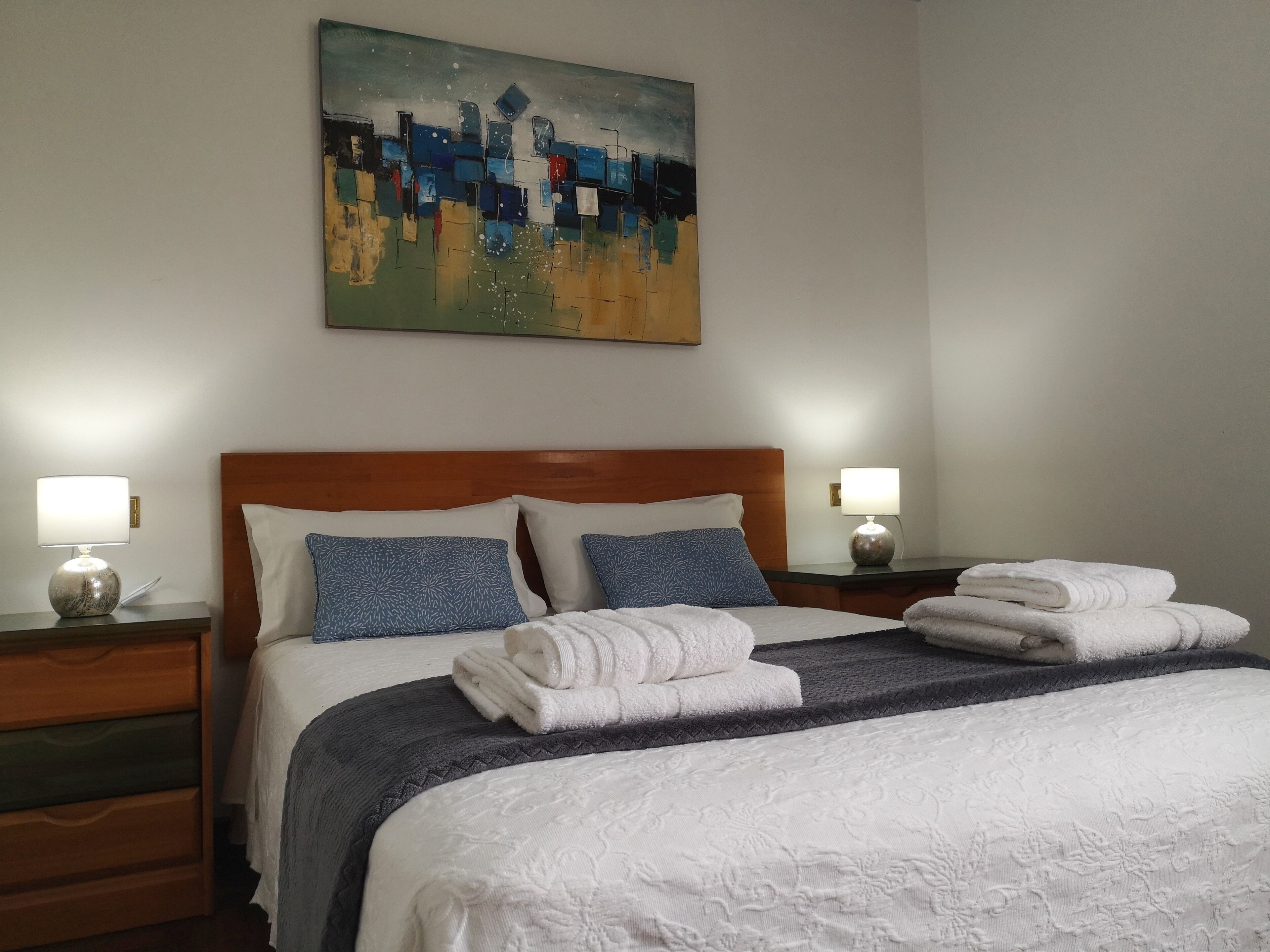 Una cama bien hecha con sábanas blancas y almohadas con motivos azules, un cabecero de madera y un cuadro en la pared superior.