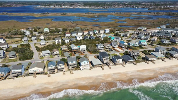 An aerial view of a coastal residential area with houses along the beach, a sandy shoreline, and a marshy area with water bodies in the background. - Your Ultimate  Retreat – Steps from Beach with PVT Heated Pool, Slide & Hot Tub! (Surf City)