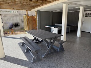 Un patio al aire libre con mesa y bancos de madera, una parrilla de barbacoa y un área cubierta con una puerta blanca.