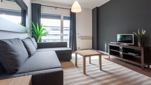 Uma sala de estar moderna com um sofá cinzento, uma mesa de centro de madeira, uma TV sobre um móvel escuro de madeira e uma grande janela com cortinas.