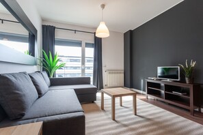 Un salon moderne avec un canapé gris, une table basse en bois, un téléviseur posé sur un meuble en bois foncé et une grande fenêtre avec des rideaux.