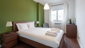 Cama bem arrumada com lençóis e toalhas brancas, abajur, mesa de cabeceira, pequena mesa com luminária e radiador.