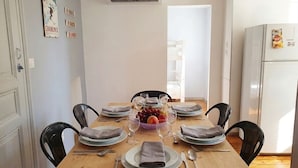 Un coin repas avec une table en bois dressée pour quatre personnes, des chaises, un panier de fruits et un réfrigérateur blanc.