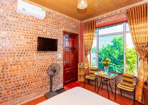 A room with brick walls, a TV mounted on the wall, a fan, a table with chairs, and a window with curtains. - Bai Dinh Eco Homestay (Gia Vien)